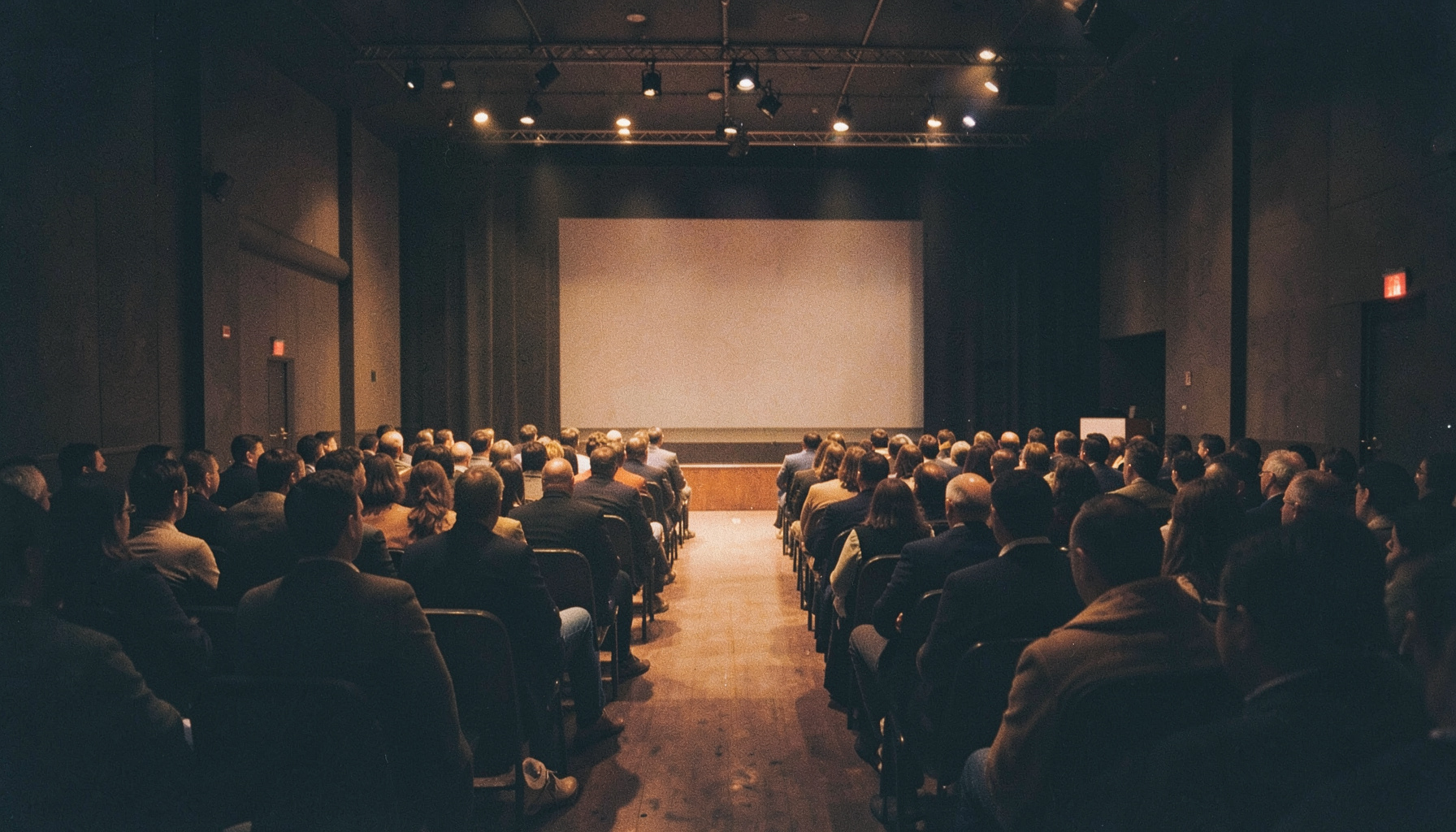 A dimly lit conference hall with rows of attendees facing a stage, warm amber and deep navy tones, soft film grain.