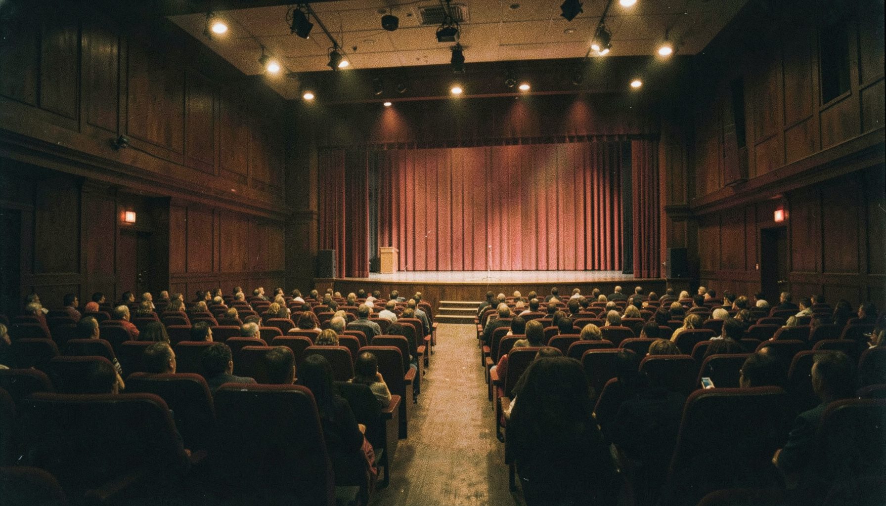 A wide conference hall with rows of seating and a stage lit by warm amber spotlights, deep red curtains framing the speaker area, dark wood panelling, and a hushed anticipatory atmosphere.